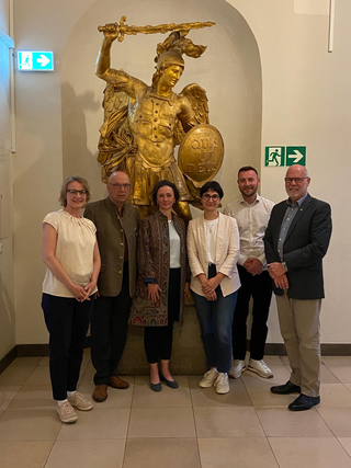 Sechs Personen (vier Frauen, zwei Männer) posieren vor einer großen goldenen Statue des Erzengels Michael mit Schwert und Schild.