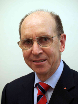 Nahaufnahme eines älteren Mannes mit Brille, dunklem Anzug, hellblauem Hemd und gestreifter Krawatte, leicht lächelnd.