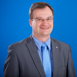 Professionelles Porträt eines lächelnden Mannes in grauem Anzug, blauem Hemd und blauer Krawatte.