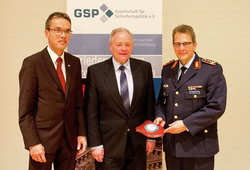 Drei Männer, darunter ein Soldat in Uniform, der eine rote Plakette hält, stehen vor einem Banner der GSP.
