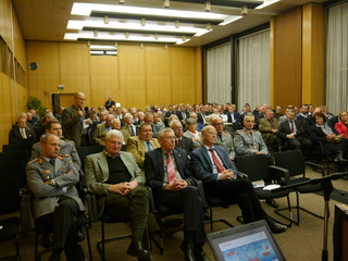 Ein Mann spricht vor einem großen Publikum von Männern, teils in Militäruniform, in einem Konferenzraum mit Holzvertäfelung.