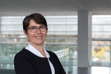 Lächelnde Frau mit Brille und kurzer dunkler Haarfrisur in schwarzer Jacke vor einem modernen Gebäude mit großen Fenstern.