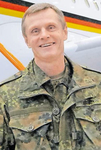 Lächelnder Mann in deutscher Flecktarn-Uniform, mit Flugzeug und deutscher Flagge im Hintergrund.