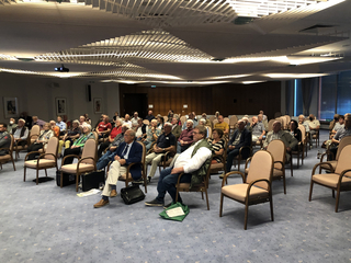 Ein Publikum aus Erwachsenen und Senioren sitzt in einem Konferenzraum mit Projektor und besonderer Deckenbeleuchtung.