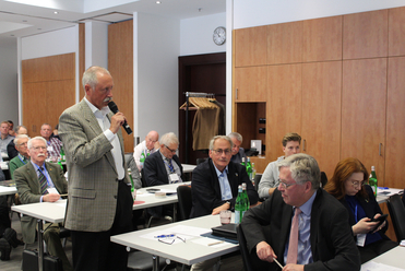 Ein Mann spricht ins Mikrofon bei einer Konferenz mit einem Publikum aus verschiedenen Personen an Tischen sitzend.