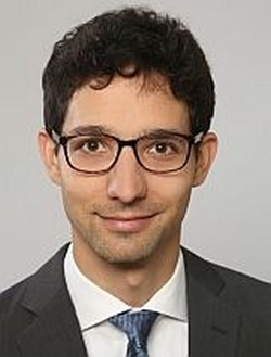 Lachender junger Mann mit dunklem Lockenhaar, Brille, dunklem Anzug und blauer Krawatte vor grauem Hintergrund.