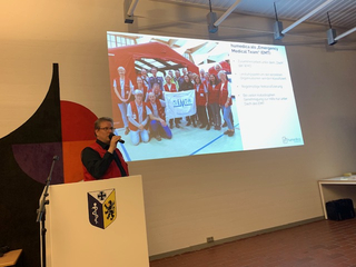 Ein Mann in roter Weste spricht an einem Rednerpult. Eine Leinwand zeigt Humedica-Freiwillige in roten Westen und ein rotes Zelt.