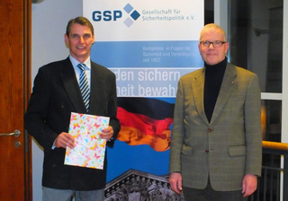 Zwei Männer lächeln vor einem GSP-Banner mit deutscher Flagge. Einer hält eine bunte Mappe mit Schmetterlingsmuster.