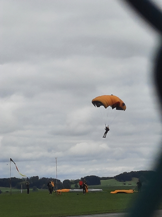 Ein Fallschirmspringer mit orangefarbenem Fallschirm nähert sich einer Landezone an einem bewölkten Tag, mit Bodenpersonal sichtbar.