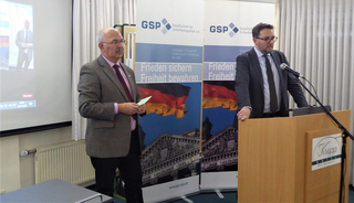 Zwei Männer bei einer GSP-Veranstaltung, einer am Rednerpult, mit Bannern der deutschen Flagge und des Reichstagsgebäudes.