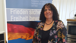 Lächelnde Frau mit dunklen Locken in Blumenjacke vor einem Banner mit deutscher Flagge und dem Text 'Frieden sichern Freiheit bewahren'.