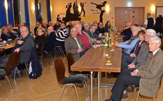 Eine große Gruppe älterer Erwachsener bei einem gesellschaftlichen Treffen oder Event in einer Halle mit Jazz-Wandkunst.