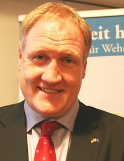 Porträt des deutschen Politikers Norbert Röttgen, lächelnd in dunklem Anzug und roter Krawatte mit Anstecknadel.