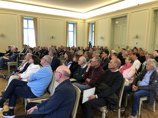 Ein diverses Publikum von Erwachsenen sitzt aufmerksam in einem eleganten, hellen Saal während einer Präsentation.