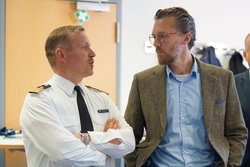 Ein Mann in weißem Uniformhemd mit Schulterklappen und Namensschild „Dr. Jentzsch“ spricht mit einem bärtigen Mann in Sakko und Brille.