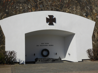 Deutsches Kriegsdenkmal mit Eisernem Kreuz, liegender Figur und Text 'Den Toten des Deutschen Heeres' unter einem weißen Bogen.