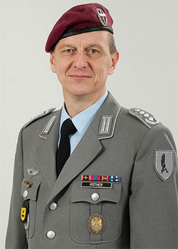 Deutscher Offizier ROTHER in grauer Uniform und bordeauxrotem Fallschirmjägerbarett, leicht lächelnd.