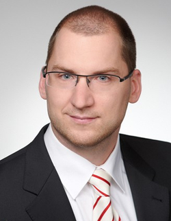 Professionelles Porträt eines lächelnden Mannes mit Brille, kurzem Haar, Anzug und gestreifter Krawatte.