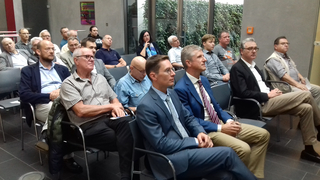 Eine vielfältige Gruppe von Männern und einer Frau sitzt aufmerksam in einem Konferenzraum und blickt auf eine Präsentation oder einen Sprecher.