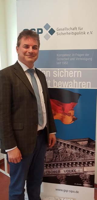 Lächelnder Mann im Anzug vor GSP-Banner mit deutscher Flagge und Reichstagsgebäude, für Sicherheitspolitik werbend.
