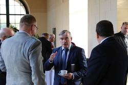 Mann im Anzug gestikuliert beim Gespräch mit anderen Teilnehmern bei einer Konferenz, Tasse in der Hand.
