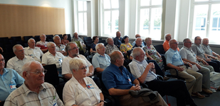 Teilnehmer, hauptsächlich Senioren, sitzen in Reihen während einer Konferenz oder eines Meetings in einem hellen Raum mit Fenstern.