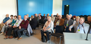 Eine Gruppe älterer Menschen sitzt auf Stühlen in einem Tagungsraum mit blauen Wänden, einige tragen Namensschilder.