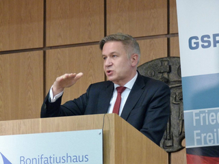 Mann im Anzug spricht an einem Holzpult und gestikuliert mit der Hand. 'Bonifatiushaus'-Logo und Banner im Hintergrund.