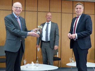 Drei Männer in Anzügen in einem Besprechungsraum. Der linke Mann lächelt und überreicht eine kleine Geschenkflasche.