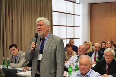Älterer Mann mit grauem Haar und Bart spricht in ein Mikrofon bei einer Konferenz, im Hintergrund weitere Zuhörer.