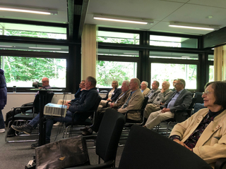 Zuhörer, meist ältere Erwachsene, sitzen aufmerksam in einem Konferenzraum mit Blick auf grüne Bäume und verfolgen eine Präsentation.