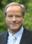 Lächelndes Porträt eines Mannes mit hellem Haar, blauen Augen, blauem Hemd und gestreifter Krawatte, vor grünem Hintergrund.