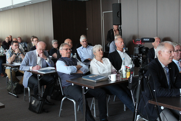 Eine vielfältige Gruppe von Erwachsenen nimmt an einer Konferenz oder einem Seminar teil, sitzt an Tischen mit einer Kamera im Vordergrund.