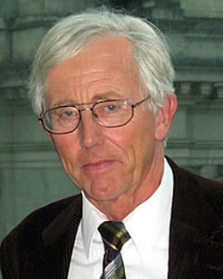 Nahaufnahme eines älteren Mannes mit weißen Haaren und Brille, der eine gemusterte Krawatte und ein Sakko trägt.