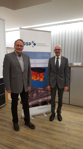 Zwei Männer lächeln vor einem Banner der Gesellschaft für Sicherheitspolitik mit Reichstagsgebäude und deutscher Flagge.