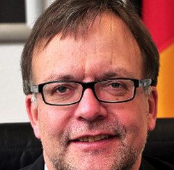 Nahaufnahme eines Mannes mittleren Alters mit Brille und leichtem Lächeln, mit der deutschen Flagge unscharf im Hintergrund.