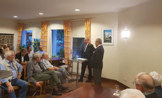 Eine Gruppe von Menschen, hauptsächlich Männer, nimmt an einer Indoor-Versammlung teil. Zwei Männer in Anzügen unterhalten sich vorne stehend.