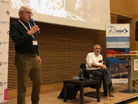 Ein Mann mit Brille spricht in ein Mikrofon bei einer Konferenz, mit Veranstaltungsbannern und einem Zuhörer im Hintergrund.
