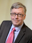 Lächelnder Mann mittleren Alters mit grauen Haaren und Brille, in dunklem Anzug, hellblauem Hemd und rosa Krawatte.