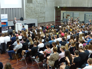 Ein Redner spricht vor einem großen Publikum junger Leute in einem Hörsaal mit Betonwänden und einem Banner.