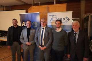 Fünf Männer in Anzügen und Business-Casual-Kleidung stehen vor Bannern, eines feiert das '70 GSP'-Jubiläum.
