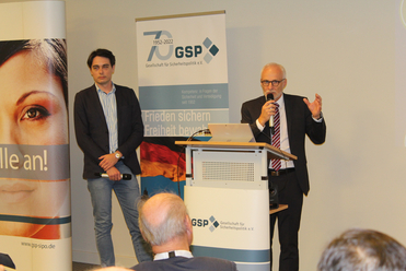 Zwei Männer, einer spricht am Rednerpult, der andere steht daneben, bei einer GSP-Sicherheitspolitik-Veranstaltung.