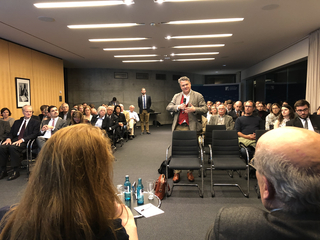 Ein Redner spricht vor einem Publikum bei einem Vortrag oder einer Konferenz, mit dem Logo der Konrad-Adenauer-Stiftung im Hintergrund.