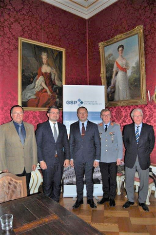 Fünf Männer, einer in Uniform, vor einem GSP-Banner und historischen Gemälden in einem roten Raum stehend.