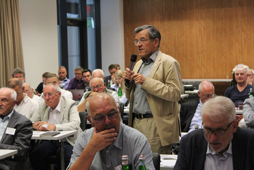 Ein Mann mit Brille und beigem Sakko spricht in ein Mikrofon bei einem Meeting vor sitzendem Publikum.