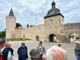 Eine Gruppe Touristen hört einem Führer auf einem Kopfsteinpflasterplatz zu, vor einer alten Stadtmauer mit Turm und Torhaus.