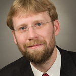 Kopfportrait eines lächelnden Mannes mit blonden Haaren, rotem Bart, blauen Augen und randloser Brille, Anzug und roter Krawatte.