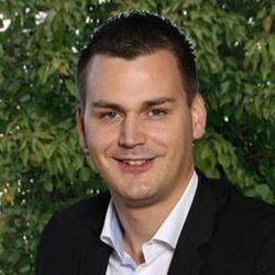 Ein lächelnder Mann mit kurzen dunklen Haaren in dunklem Anzug und weißem Hemd, vor einem unscharfen grünen Hintergrund.