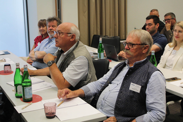 Eine Gruppe von Personen nimmt an einer Konferenz oder Besprechung teil, sitzend an Tischen mit Wasserflaschen, Papieren und Laptops.