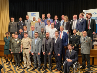 Vielfältige Gruppe, teils in Militäruniform, teils in Zivil, darunter ein Mann im Rollstuhl, posiert für ein Gruppenfoto.